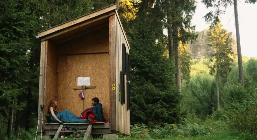 Zwei Personen entspannen sich in einer einfachen Holzhütte, bereit für wild camping. Umgeben von hohen Bäumen und üppigem Grün.
