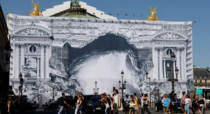 Des passants traversent devant l'Opéra Garnier, dont la façade est recouverte d'une bâche monumentale simulant des travaux et une arche rocheuse.
