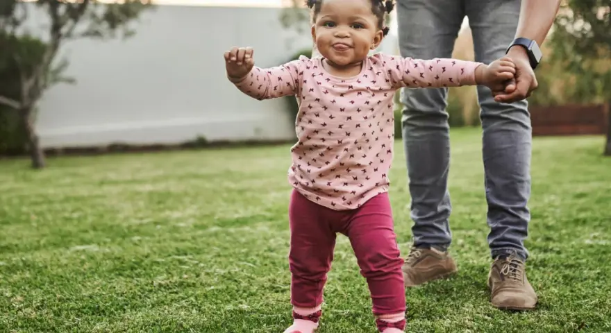 Ein kleines Mädchen mit lustigen Zöpfen macht seine ersten Schritte im Gras, gehalten von einer erwachsenen Hand. Wann laufen Babys? Heute ist ein guter Tag dafür!