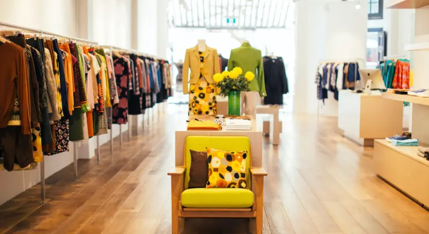 Interior de un showroom de ropa con percheros llenos de prendas coloridas y un sillón verde con cojines estampados.
