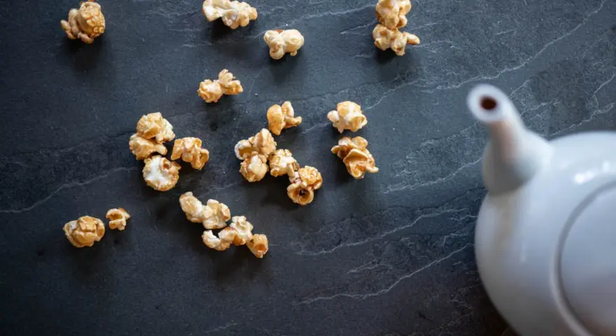 Pop corn caramel éparpillé sur une surface sombre, avec une théière blanche en arrière-plan.