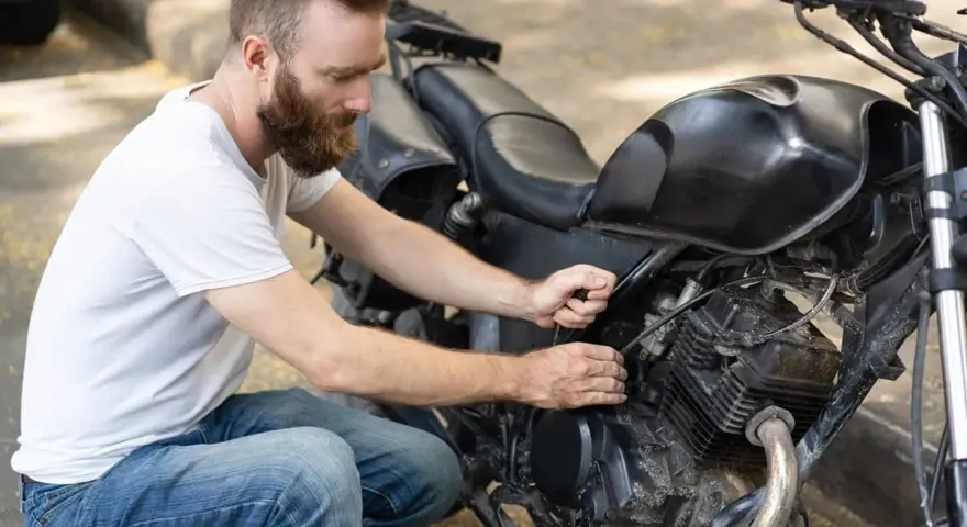 Uomo con barba lavora sul motore di una moto. Controlla i cavi, forse per il tempo di ricarica batteria moto 12v.