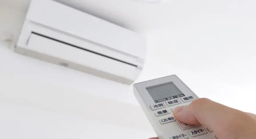 Mano ajustando el **timer on aire acondicionado** con el control remoto. El aire acondicionado blanco está montado en la pared.