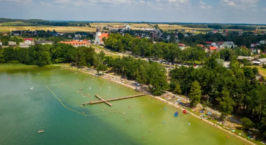 Letnie popołudnie nad jeziorem wielewskim. Tłumy plażowiczów, kąpielisko z pomostem i urokliwa miejscowość w tle.