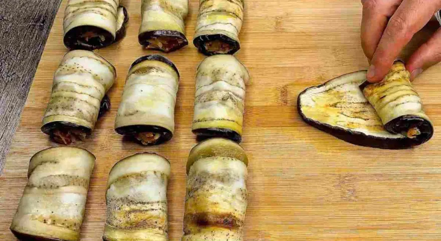 Nove involtini di melanzane pronti, uno viene arrotolato su una fetta di melanzana grigliata.