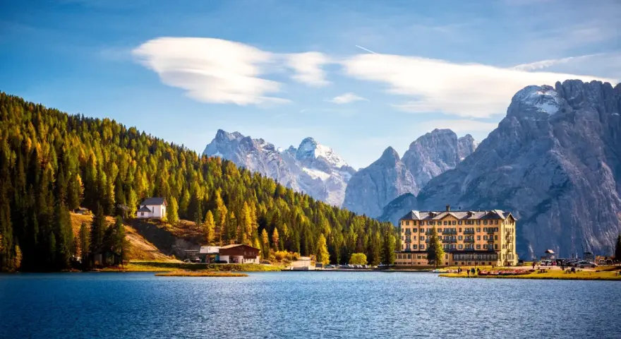 Laghi del Veneto in moto: un hotel giallo e una chiesetta bianca su un pendio boscoso, con montagne innevate sullo sfondo.