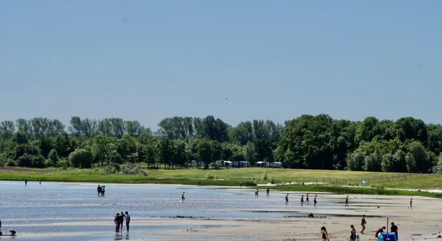 Menschen genießen einen sonnigen Tag am Strandbad Eldena. Einige baden im flachen Wasser, andere spazieren am Ufer entlang.