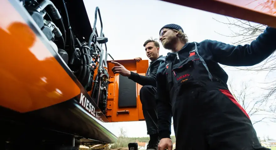 Dwóch mechaników w kombinezonach przeprowadza konserwację maszyn budowlanych. Jeden z nich wskazuje na silnik.