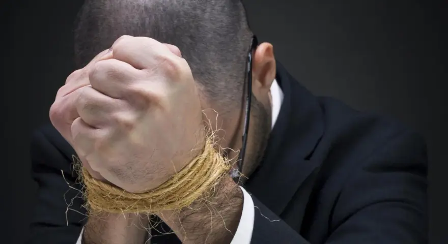 Mężczyzna w garniturze z rękami związanymi sznurkiem, pochylony.