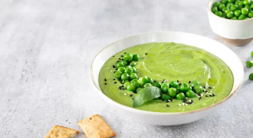 Cremoso hummus di piselli guarnito con piselli freschi, basilico e semi di sesamo, servito con cracker croccanti.