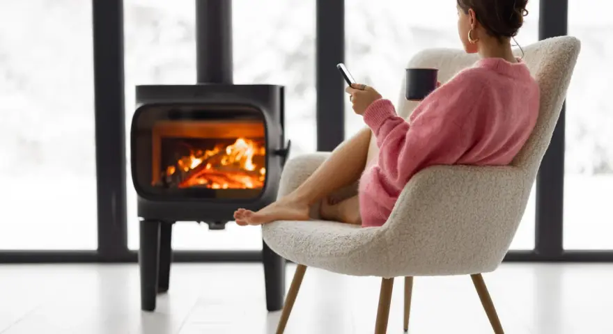 Femme assise près d'un poêle à bois, tenant une tasse et un téléphone.