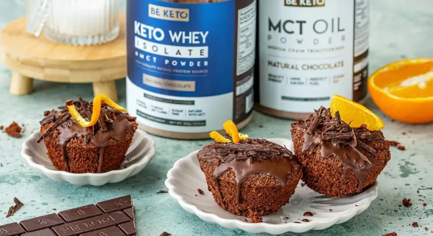 Pyszne keto muffiny z mąki migdałowej, polane czekoladą i ozdobione skórką pomarańczową. Obok stoją puszki z odżywką białkową i olejem MCT.