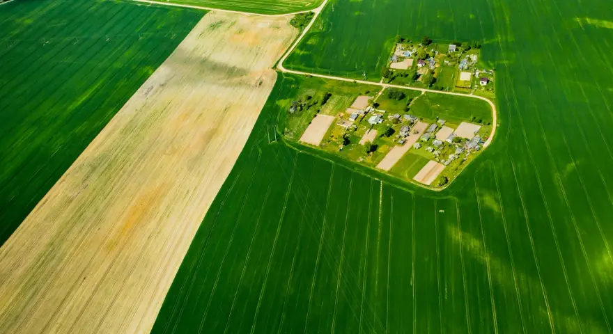 Widok z lotu ptaka na pola uprawne i wieś. Różne odcienie zieleni i żółcieni sugerują różne rodzaje gleby, co może pomóc w zrozumieniu, jak sprawdzić klasę gruntu.