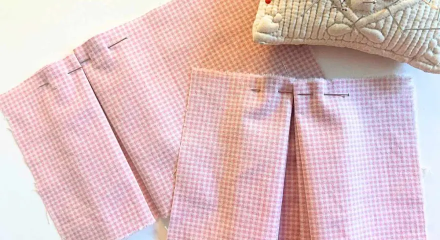 Tissu rose à carreaux avec des plis épinglés, montrant comment faire des plis en couture avec une fourchette. Coussin à épingles en arrière-plan.