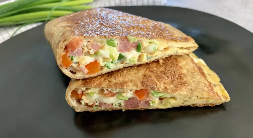 Keto tortilla przepis: złocisty placek z jajek, szynki, pomidorów i szczypiorku, złożony na pół, na czarnym talerzu.