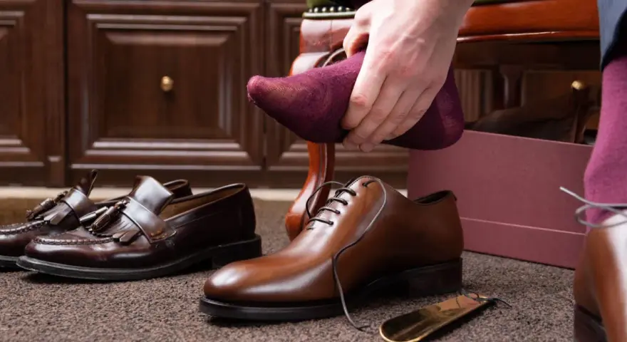 Hombre se pone calcetín morado para ablandar zapatos con crema hidratante. Zapatos de vestir y mocasines en el suelo.