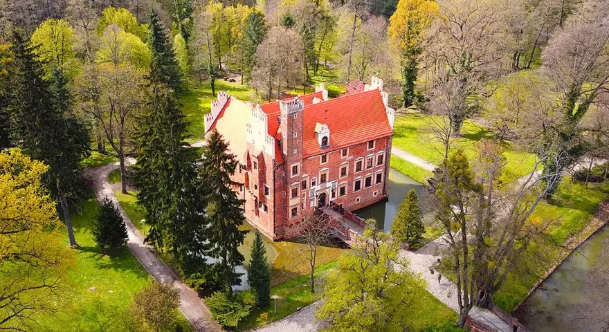 Zamek na wodzie w Wojnowicach otoczony zielenią i drzewami. Czerwona bryła zamku z charakterystycznym dachem wznosi się nad fosą.