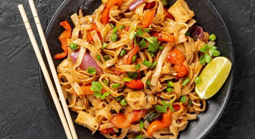 Pyszny pad thai z warzywami: makaron ryżowy, tofu, papryka, cebula i groszek. Podany z limonką i pałeczkami.