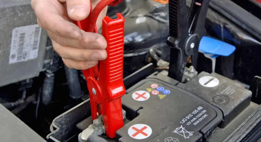 Hand klemmt rote Überbrückungsklemme an die Autobatterie, um Plus und Minus zu verbinden.