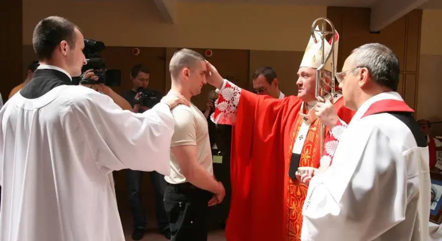Biskup udziela sakramentu bierzmowania młodemu mężczyźnie. W tle widać osoby z kamerami.