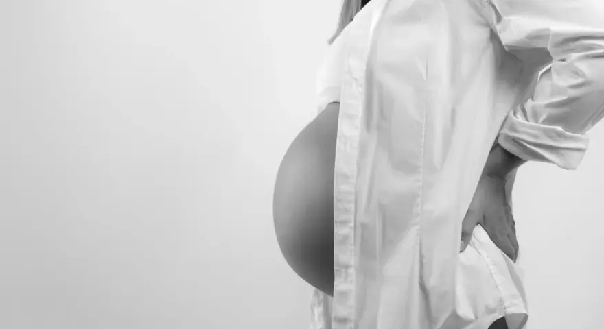 Brzuszek w 7 miesiącu ciąży, otulony białą koszulą. Delikatne światło podkreśla krągłości.