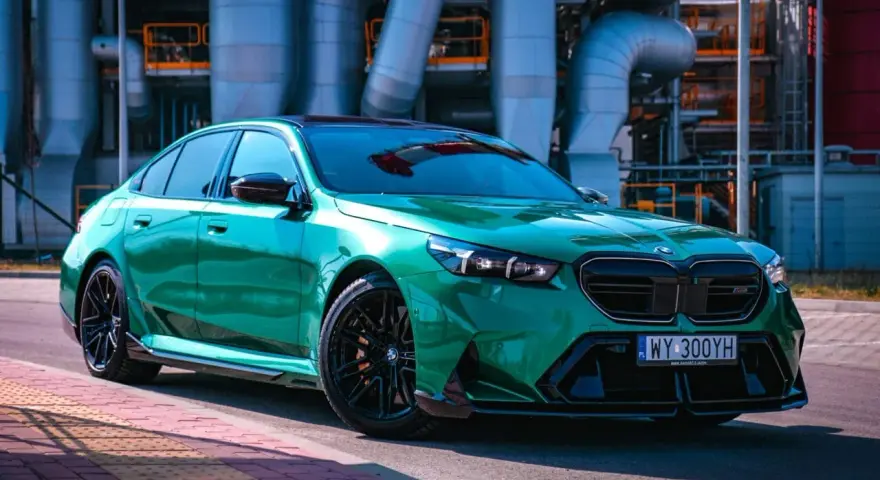 Błyszczące, zielone BMW M5 G90 z czarnymi felgami stoi na tle przemysłowych rur.