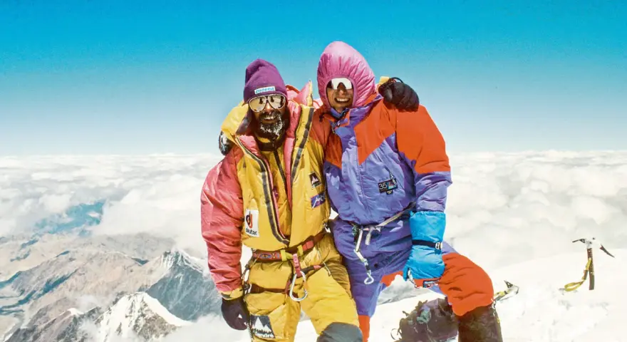 Zwei Bergsteiger auf dem Gipfel, die sich umarmen. Sie tragen bunte Daunenanzüge und Mützen. Die Frage "wie hoch ist der Mount Everest" wird hier beantwortet: 8848 Meter.