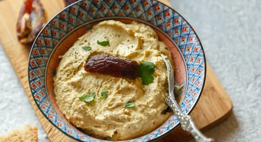 Cremiger Dattel Dip in einer bunten Schale, garniert mit einer Dattel und Petersilie. Dazu Brot und weitere Datteln auf einem Holzbrett.