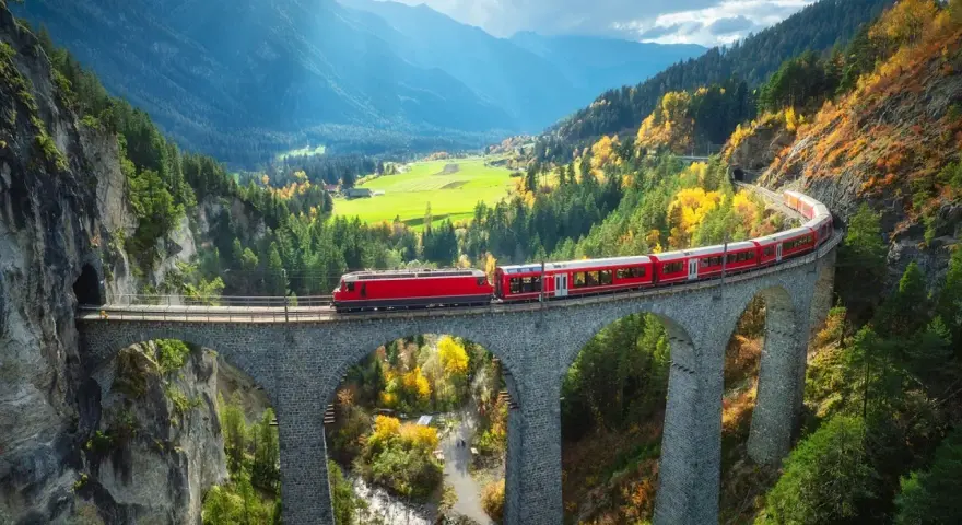 Czerwony pociąg Bernina Express przejeżdża przez kamienny wiadukt w górach jesienią.