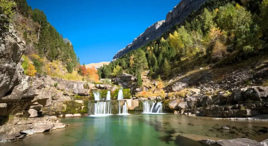 Cascada en una zona de senderismo, con aguas cristalinas y árboles de otoño en las laderas rocosas.