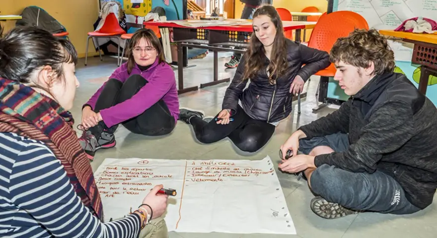 Quatre jeunes discutent autour d'un papier, participant à un mouvement d'éducation populaire. Ils brainstorment des idées pour améliorer une activité.