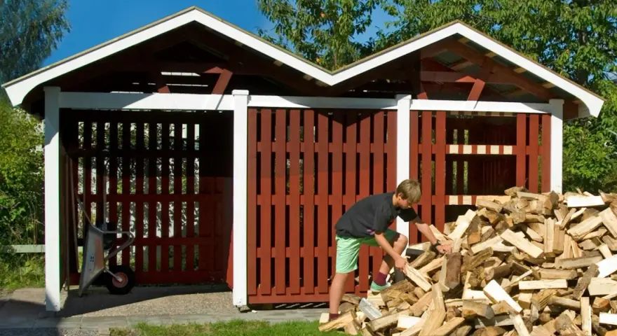 Pojke hjälper till att bygga redskapsbod genom att stapla ved. En skottkärra står redo i den tomma boden.