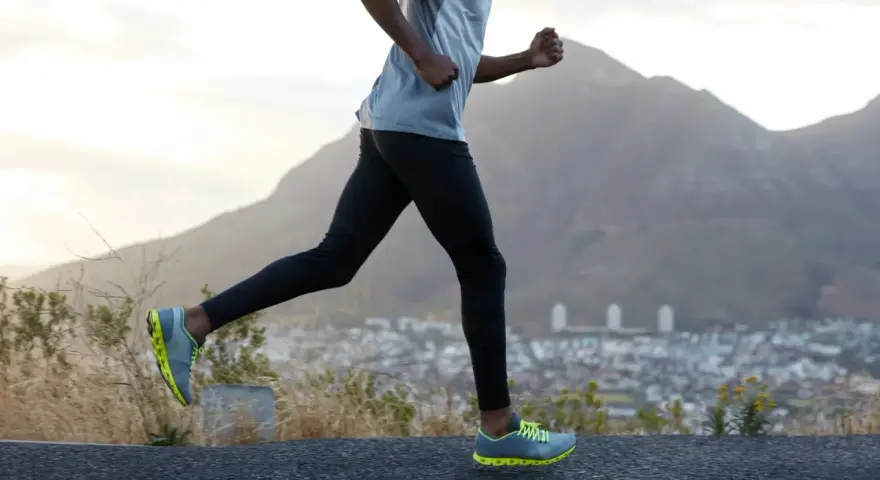 Un homme en tenue de sport fait du cardio training en courant sur une route de montagne, avec une ville en contrebas et des montagnes en arrière-plan.