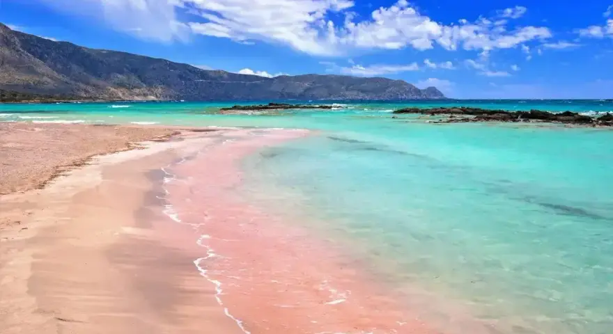 Różowy piasek plaży Elafonisi w Grecji, turkusowa woda i góry w tle.
