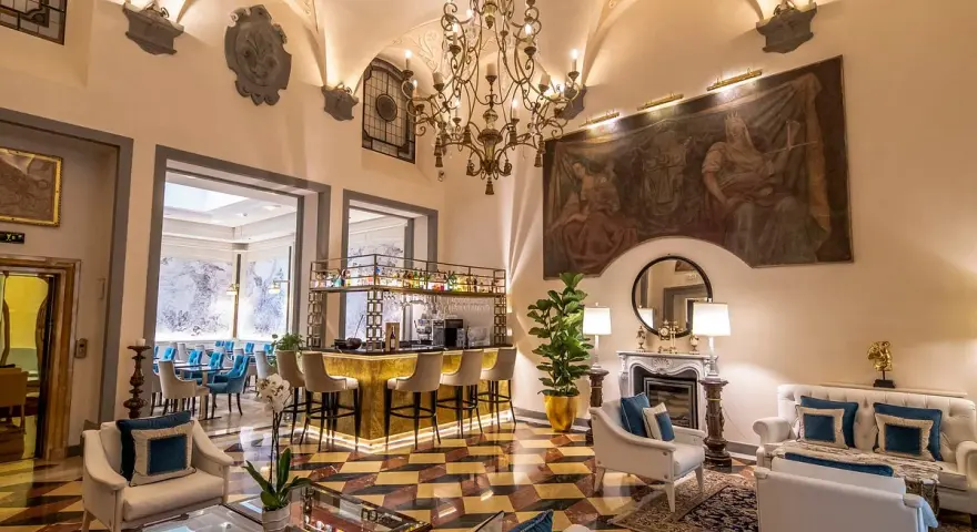 Elegant hotel lounge with a bar, fireplace, ornate chandelier, and artwork.