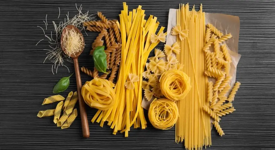 Różnorodne rodzaje makaronu, od cienkich nitek po tagliatelle i fusilli, ułożone na ciemnym tle. Wartości odżywcze makaronu zależą od jego składu.