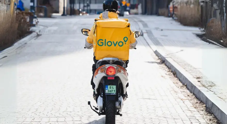Kurier na skuterze z żółtym plecakiem Glovo jedzie ulicą.