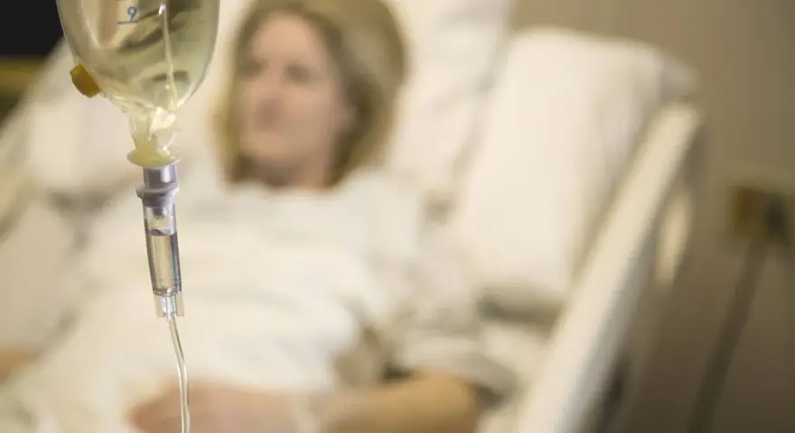 Infusion vorbereiten: Eine Frau liegt im Krankenhausbett, während eine Infusionstropfflasche und ein Tropfschlauch im Vordergrund zu sehen sind.