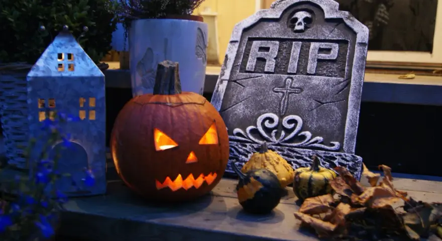 En läskig pumpa lyser upp med sitt ansikte, bredvid en gravsten och höstlöven. Perfekt för en kuslig bål halloween-kväll.