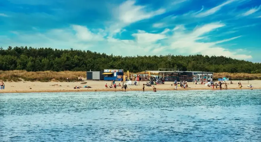 Słoneczny dzień na plaży, ludzie odpoczywają, a w tle widać budynki i las. Idealne miejsce na kemping na Helu.