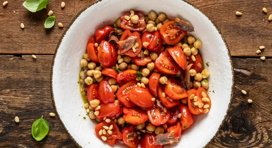 Insalata di ceci con pomodorini, acciughe e pinoli, guarnita con basilico fresco su un tavolo di legno rustico.