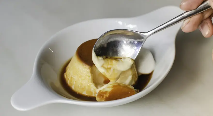 Una cuchara saca un trozo cremoso de flan de cuajada, bañado en caramelo líquido.