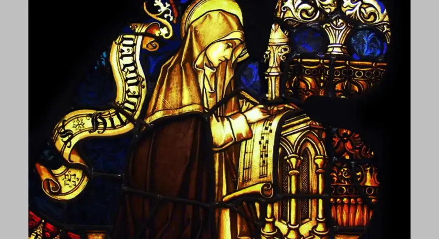 Stained glass depicts a cloaked figure, likely the head of a monastery, diligently writing on a scroll.