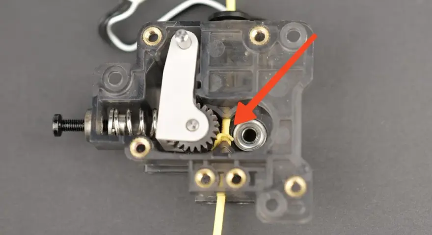 Mechanizm ekstrudera drukarki 3D, gdzie żółty filament wchodzi do mechanizmu. Strzałka wskazuje miejsce, które może wymagać czyszczenia, aby dowiedzieć się, jak wyczyścić dysze drukarki.
