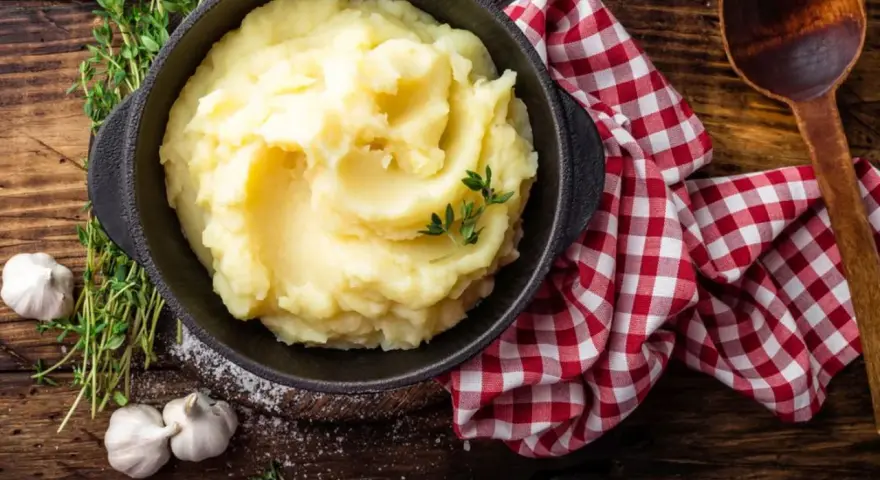 Una ciotola di cremoso purè di patate, guarnito con timo fresco, accanto ad aglio e un tovagliolo a quadri rossi.