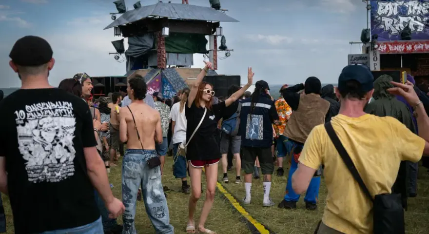 Une foule danse sous la pluie à un festival, une femme au centre lève les bras. On aperçoit une scène et des bannières, peut-être un dj français anime l'événement.