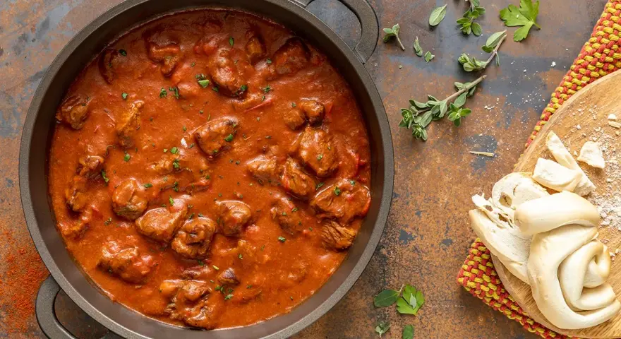 Un saporito gulash di manzo in una padella, accompagnato da pane fresco e erbe aromatiche.