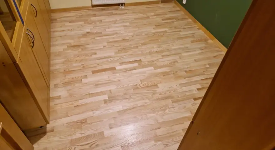 Jasny, odnowiony parkiet po cyklinowaniu, lśniący w świetle. Obok drewniane meble i zielona ściana.