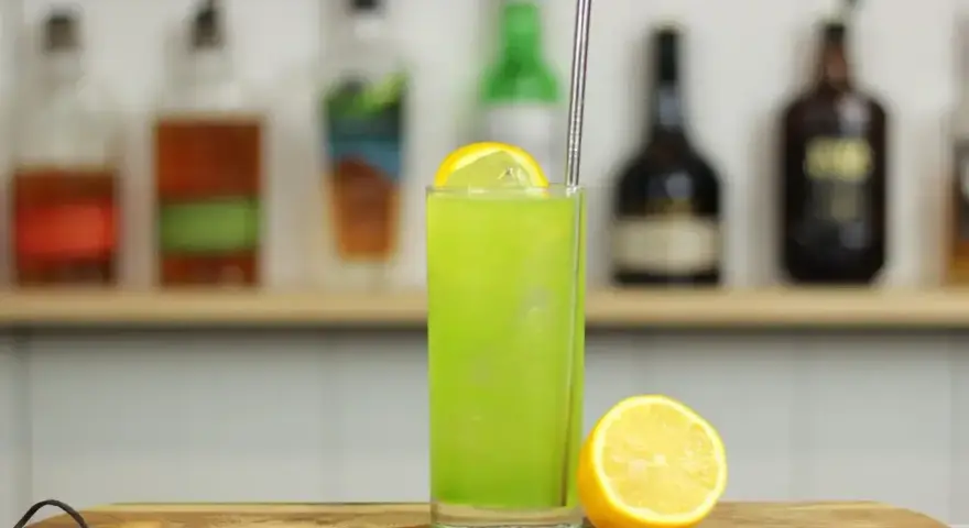 Zielony, orzeźwiający drink Tokyo Iced Tea z plasterkiem cytryny i metalową słomką, na tle półki z butelkami alkoholu.