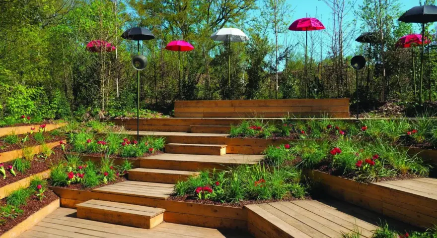 Un jardin en pente moderne avec des terrasses en bois, des parterres de fleurs rouges et des parasols colorés.